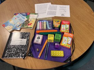 Contents of summer book bags for kindergarteners. Clockwise: worksheets, crayons, cards, diece, alphabet and math cards, book bag, composition notebook, and age appropriate books.